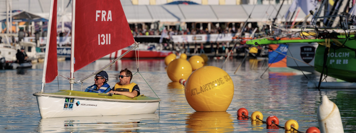 Duo de régatiers handivalide naviguant à bord d'un dériveur adapté devant les étraves de monocoques de course au large